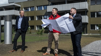 Drei Männer stehen vor einem Neubau. Zwei Männer halten einen Bauplan in der Hand.