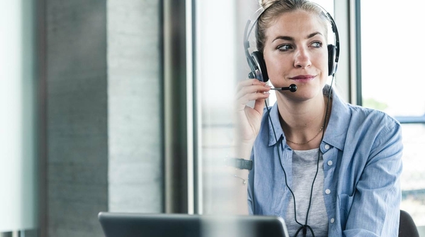 Eine Frau sitzt im Büro an ihrem Schreibtisch vor dem Laptop und telefoniert über ein Headset